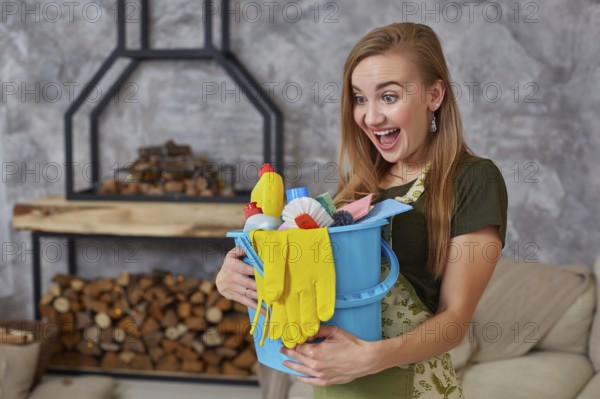 A cleaning woman is standing in living room holding a blue bucket fulfilled with chemicals and facilities for tidying up in her hand. Cleaning service