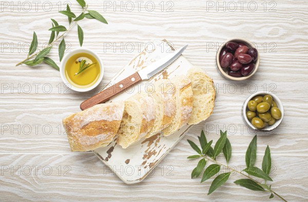 Sliced fresh ciabatta on cutting kitchen board, green and brown olives, olive oil with rosemary, olive tree branches, white wooden rustic background top view, food photography, food photography