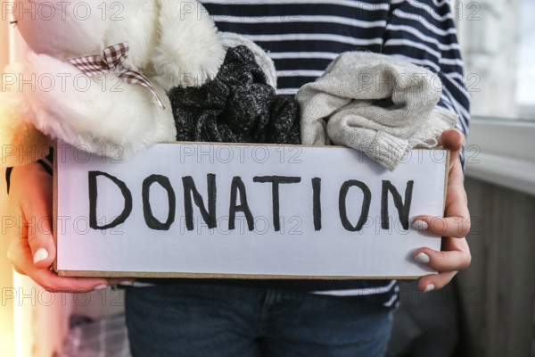 Female volunteer holding donation box with old used toys and clothes indoors. Happy Charity. Unrecognizable woman holding box with clothes in it. close-up. Clothing donation. Winter clothes in a cardboard box. Seasonal clothing