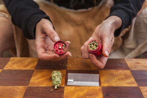 Selective focus closeup of person's hands holding a red weed filled grinder shot from above. Checkered table with rolling paper, filter and bud displayed below