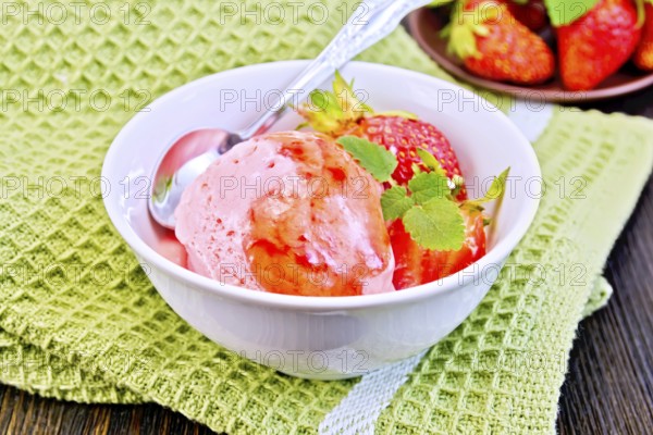 Strawberry ice cream in a white bowl with strawberries, strawberry syrup and a spoon on a napkin on a wooden boards background