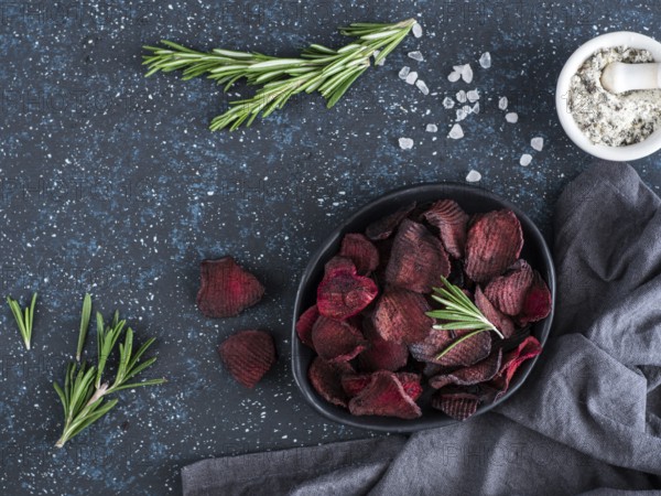Baked beet slices with coarse sea salt scented rosemary. Vegan diet food idea and recipe. Healthy homemade beetroot chips in plate and ingredients on dark blue background. Copy space. Top view