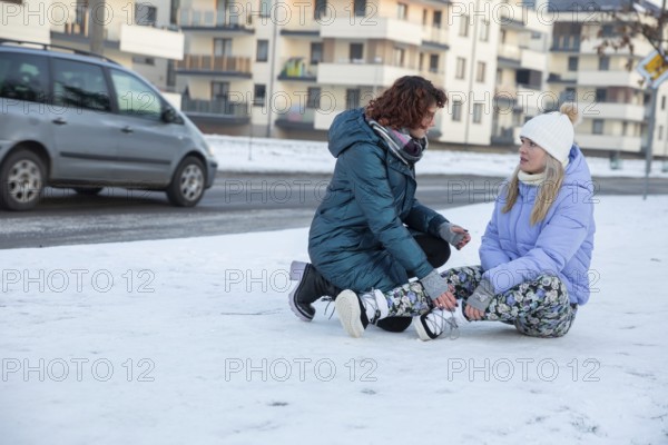 A woman who has fainted is sitting on the sidewalk. The sidewalk is covered with snow. Next to the woman crouches a passerby, who tries to help. In the background you can see a car driving down the street and an apartment block