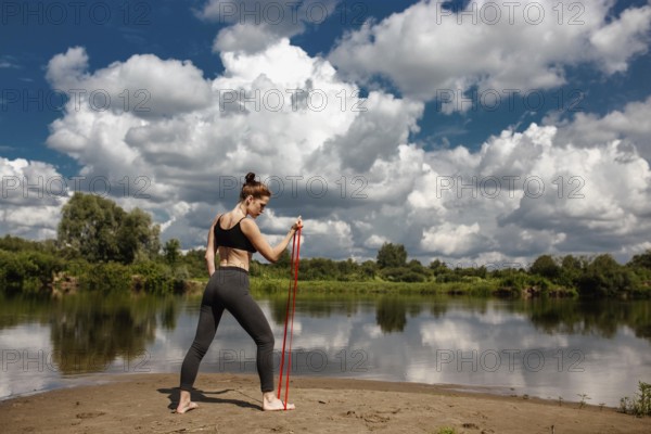 A woman exercises using a resistance band by a tranquil river, with dramatic clouds above
