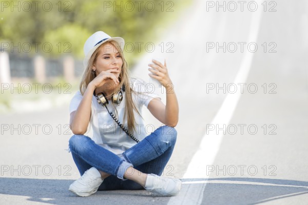 Happy young woman with vintage headphones around her neck, surfing the internet on her smartphone and sitting on a divider against the backdrop of a street
