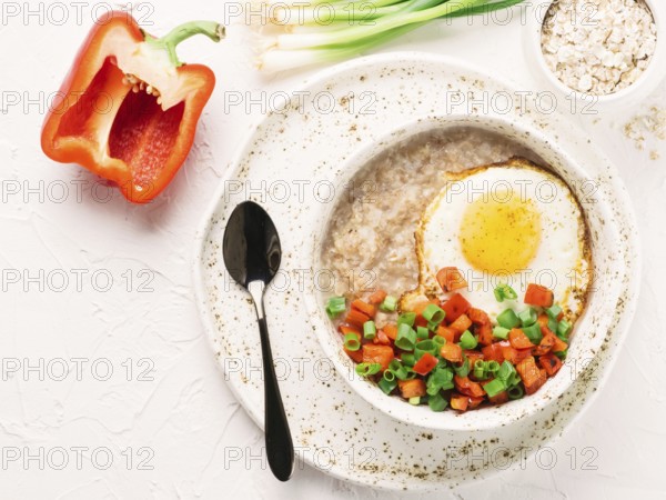 Savory oatmeal in craft trendy bowl, served with vegetables and fried egg on white concrete background. Red bell pepper and bunch onion on background. Top view or flat lay. Copy space