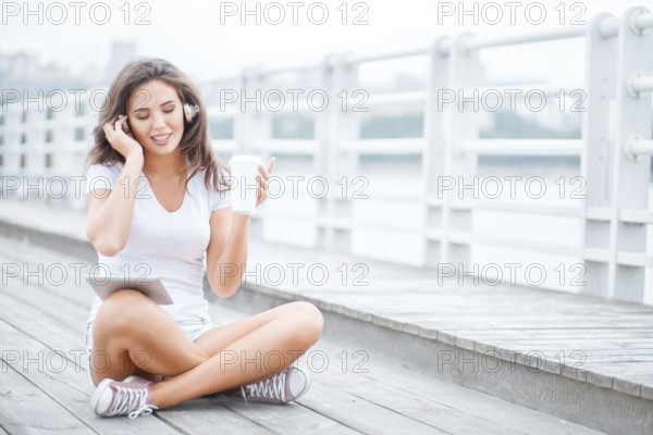 Happy young woman with music headphones and a cup of coffee to go, surfing the internet on a tablet PC, listening to music and sitting on the bridge
