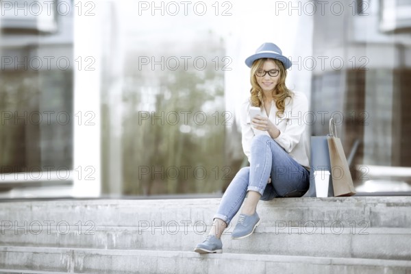 Cheerful young woman with a disposable coffee cup and shopping bags, sitting on the stairs and using her smartphone to communicate via Wi Fi Internet in a summer park