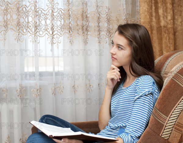A young caucasian girl dreams over a book looking thoughtfully through the window