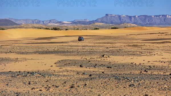 An off-road vehicle moves through a vast landscape of sand dunes and mountains, With off-road vehicles through the endless landscape of the Sahara desert in Algeria