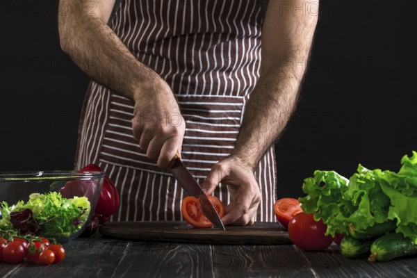 Man in a striped apron preparing salad on a wooden table. Men's hands cut the tomato to make a salad on black background. Healthy food concept