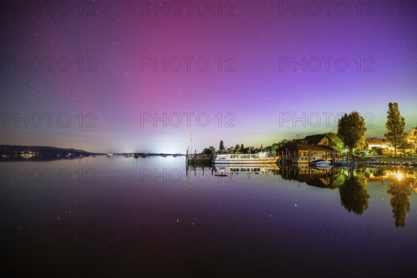 In a calm lake, the vivid northern lights are reflected next to boats and trees, harbour, Allensbach, Lake Constance, Baden-Württemberg, Germany, Europe