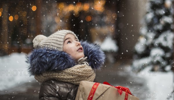 Happy girl with a gift box for Christmas catches snowflakes with his tongue on a city in winter with snow on festive market with decorations and lights. Warm clothes, knitted hat, scarf and fur