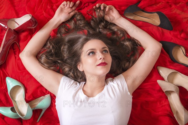 Beautiful woman with blue eyes and red lips, lays on a red fabric, smiling and surrounded by many elegant shoes in a variety of colors