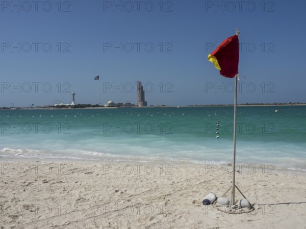 A red warning sign stands on the beach with turquoise water and a blue sky in the background, shimmering green sea with beach, parasols and sunbeds in front of a bright blue sky, Abu dhabi, Arab Emirates