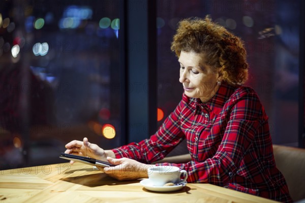 Mature woman with blond curly hair checks mail and social media notifications via tablet, smiling female holding tablet, watching video, surfing internet inside coffee shop in evening by window