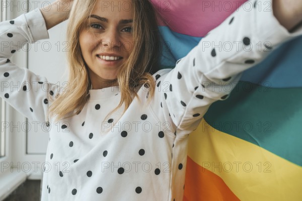 Young blonde millennial hippie woman with rainbow LGBTQ flag at home balcony. Peace and freedom the symbol of LGBT, spending and sharing loving time. Support LGBTQ community. Young lesbian woman
