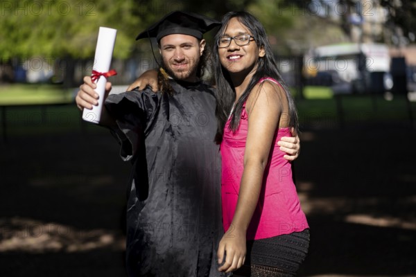 Young recently graduated man, dressed in cap and gown, with his degree in hand, celebrating with his family