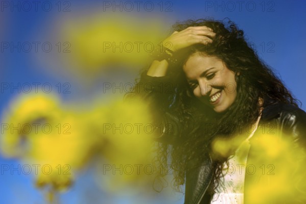 Close up portrait of a young woman holding her hand on her head and smiling in a field of yellow flowers on a sunny spring day