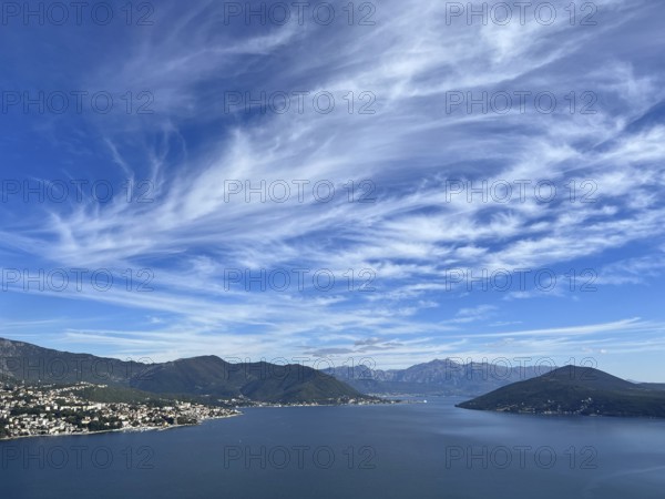 White cirrus clouds in the blue sky over a mountain range near the sea.