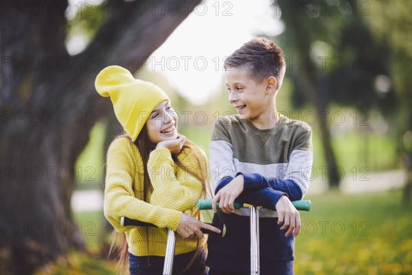 Two children caucasian boy and girl twins posing with scooters in autumn park. Happy kids riding kick scooters in public park. Kids sport. Outdoor activities brother sister teens. Happy childhood
