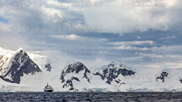 A ship in front of a snow-covered mountain range in a cold, cloudy Antarctic scenery, cruise ship in the Southern Ocean in Antarctica