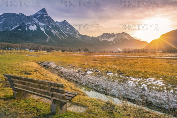 Charming winter scenery with a wooden bench on the side of a small river with a view towards the Austrian Alps, at sunset