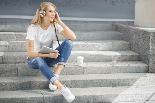 Happy young woman with vintage headphones and a cup of coffee to go, surfing the internet on a tablet PC, listening to music and sitting on a staircase in front of an urban background