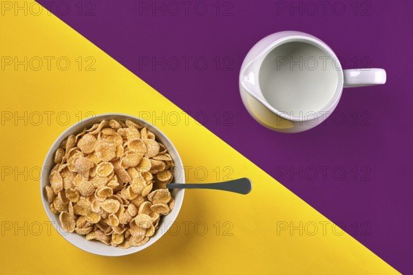 Food, healthy eating, people and diet concept. Bowl with corn flakes, jug of milk and spoon on purple and yellow background, top view. Copy space. Still life. Flat lay