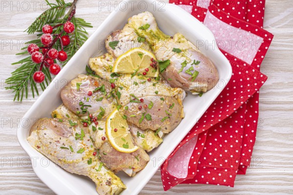 Preparing festive Christmas Eve meal with marinated chicken drumsticks. Raw chicken legs with herbs, lemon in white ceramic casserole top view on light wooden kitchen table with Christmas decoration, food photography, food photography