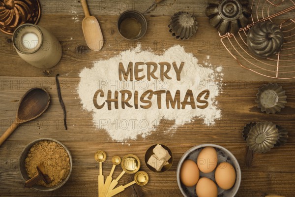 Collection of ingredients and utensils for baking on a dark wooden table with the words Merry Christmas written in flour in the centre