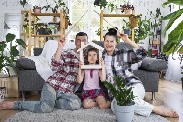 Traditional family with child are enjoying new home, sitting on the sofa and holding the roof. Mortgage, insurance and protection, buying and moving into your own home, green house with potted plant
