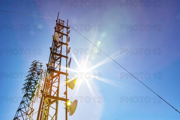 A low angle view of two mobile communication data transmitter and GPS signal towers, against a blue sky with bright sun shine and colorful lens flare