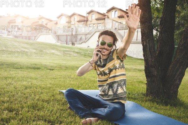 Young stylish handsome man is sitting on grass, resting and talking on the phone. waves his hand welcoming friends