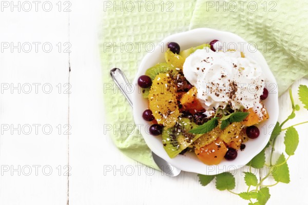 Fruit salad with orange, kiwi, cranberries and baked pumpkin, whipped cream, sprinkled with chocolate and coconut with mint in a bowl on a towel on wooden board background from above