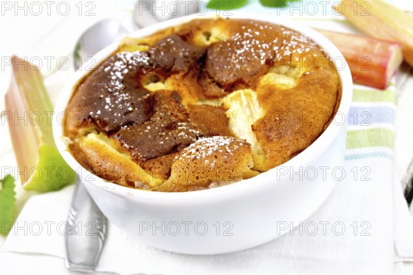 Pie klafouti with rhubarb in the bowl, mint and rhubarb stalks on a napkin against light wooden board
