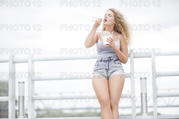 Happy young woman with long curly hair holding a lunch box and eating noodles from a Chinese snack bar with chopsticks