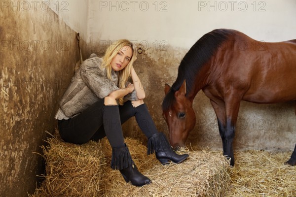 Blond woman on an indoor stable with a brown horse on straw bale