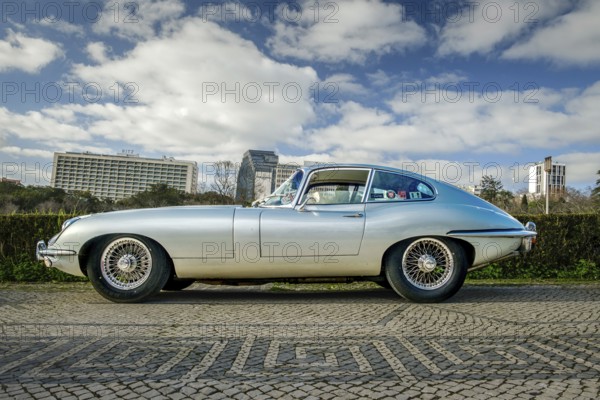 Lisbon, Portugal, Jan 20, 2024: Full side view of classic silver E-Type Jaguar Series 3 coupe car with chrome detailing parked on a cobblestone path near green park, Europe