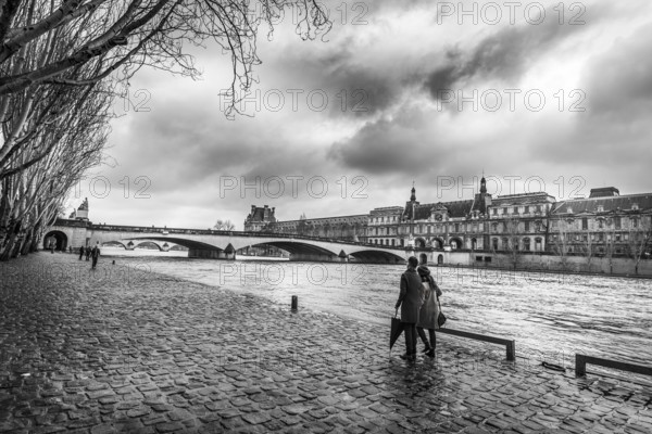 Black and white image with the Seine River, its shore where people walk, an old bridge and the Orsay Museum, under a dramatic sky, on a chilly day of February