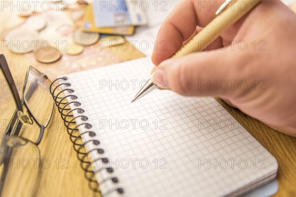 Close-up image with a blank math notebook and a man's hand holding a pen. In the background there are money, cards, bills and a pair of eyeglasses