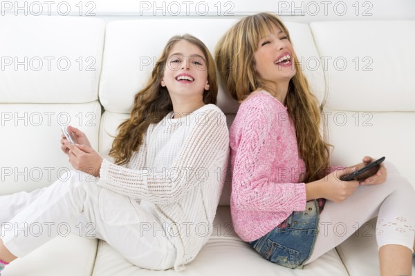 Children friends kid girls having fun playing back to back with tablet pc on white sofa