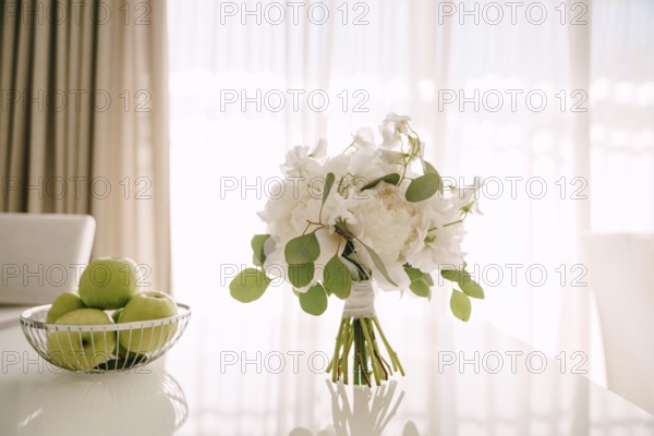 Bridal bouquet of white peonies, sweet pea and branches of eucalypt tree on a glossy white table and bowl with green apples near it.