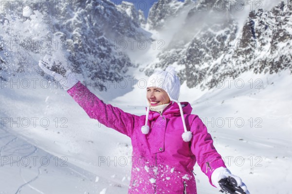 Young woman in pink ski jacket and white gloves smiling and throwing snow in the air with mountain in the background. Skalnate Pleso ski resort, Slovakia, Europe