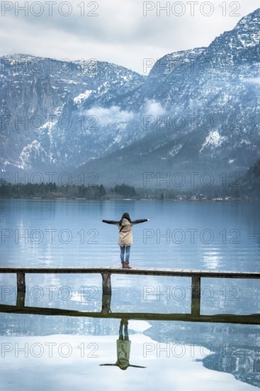 Girl with her arms open, standing on a narrow wooden deck, enjoying the view of the Austrian Alps mountains reflected in the Hallstatter lake water, in Austria