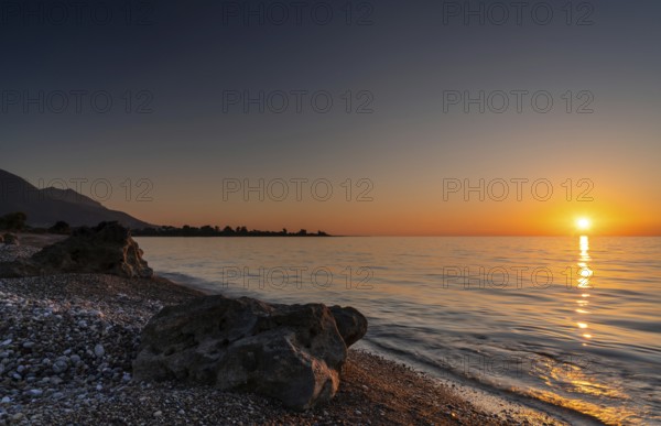 A peaceful sunset over the ocean with silhouette of mountains and pebble beach iwth rock boulders in the foreground