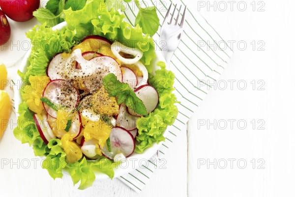 Radish, onion and orange salad with mint, vegetable oil and spices on lettuce in a plate on a towel on wooden board background from above