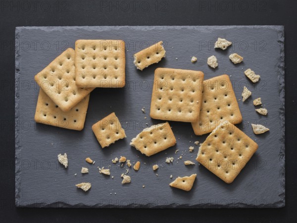 Square crackers with pieces and crumbs on slate gray background. Dry salt cracker cookies with fiber and dry spices. Top view or flat lay