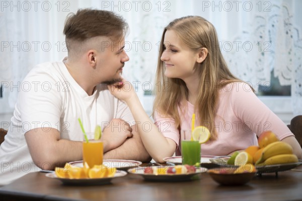 On a wooden table stand plates with various fruits. At the table sits a young couple. The man and woman are smiling at each other, and she is stroking his face with her hand