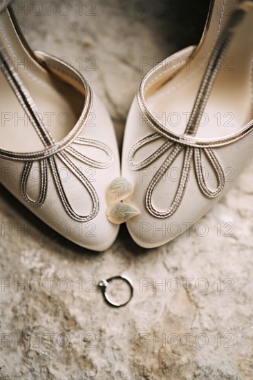 Women's earrings in the shape of leaves on the toes of the bride's white shoes with an engagement ring on a rough texture.